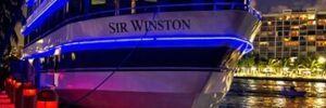 A large yacht named "Sir Winston" docked at night, illuminated with blue lights, beside a walkway with red lights.