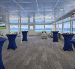 Spacious event room with blue cocktail tables, floral centerpieces, large windows, ocean views, and lounge seating.