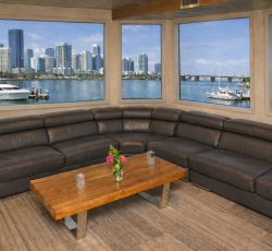 A modern lounge with a dark leather sectional sofa, wooden table, and large windows showing a city skyline and waterfront.
