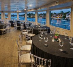Dining area on a boat with round tables, black tablecloths, and clear chairs, overlooking water and waterfront homes.