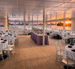 Elegant dining room on a boat set for an event, with round tables and a sunset view over the water through large windows.